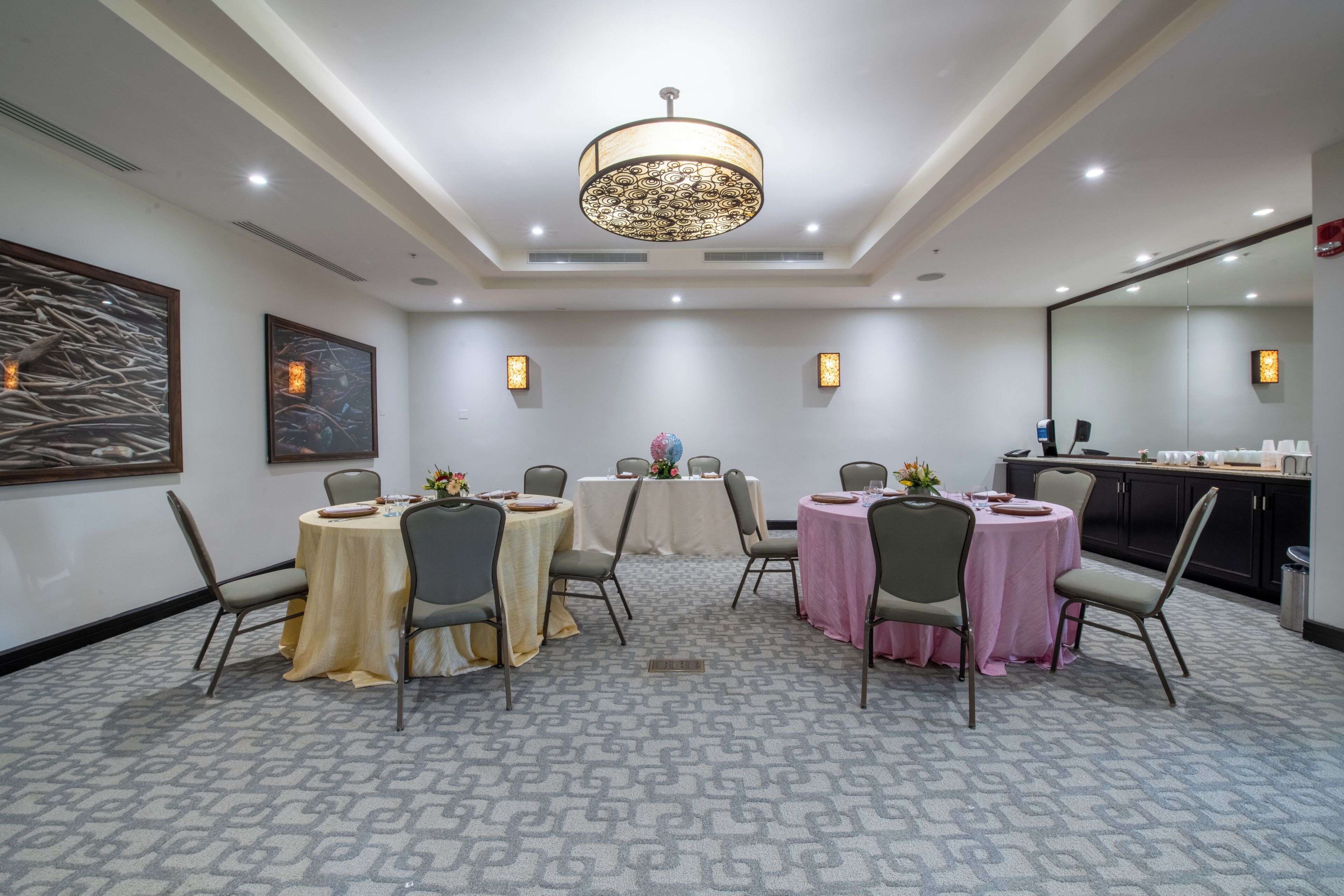 A conference room with four tables, each with a different colored tablecloth. There are chairs around the tables, and a large chandelier hangs from the ceiling. The room has a carpeted floor and a large mirror on one wall.