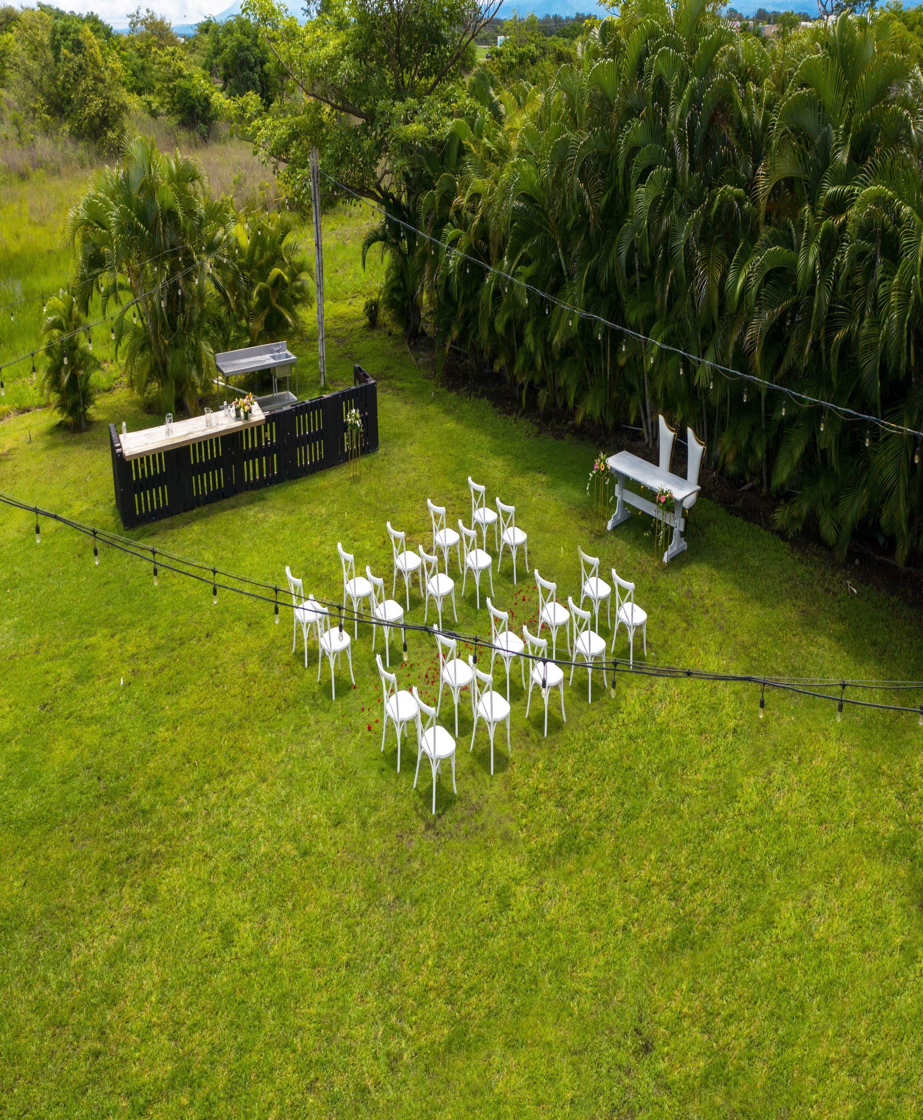 An outdoor wedding setup with white chairs arranged in a semi-circle, a wooden bench, and a black fence with a table and chairs.