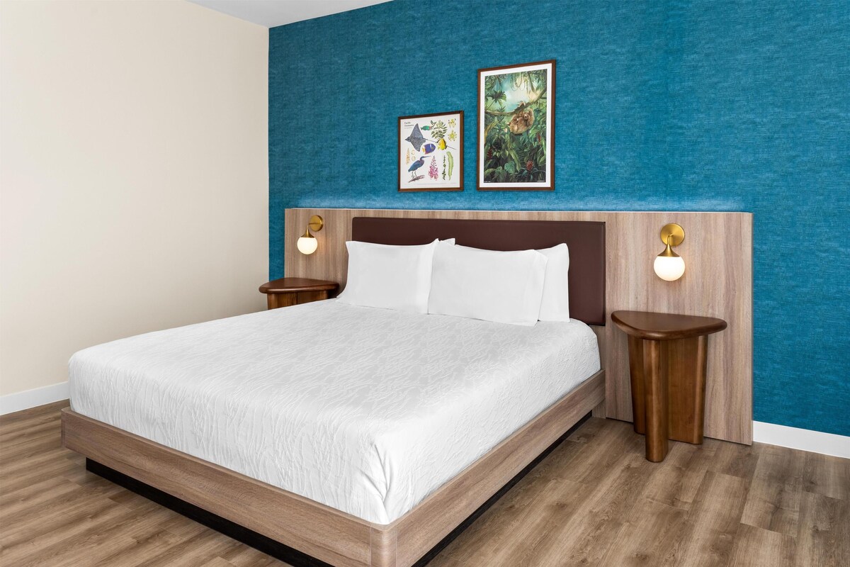 Une chambre d’hôtel avec un grand lit, une tête de lit en bois, deux tables de chevet fixées au mur et deux tableaux encadrés au-dessus du lit.