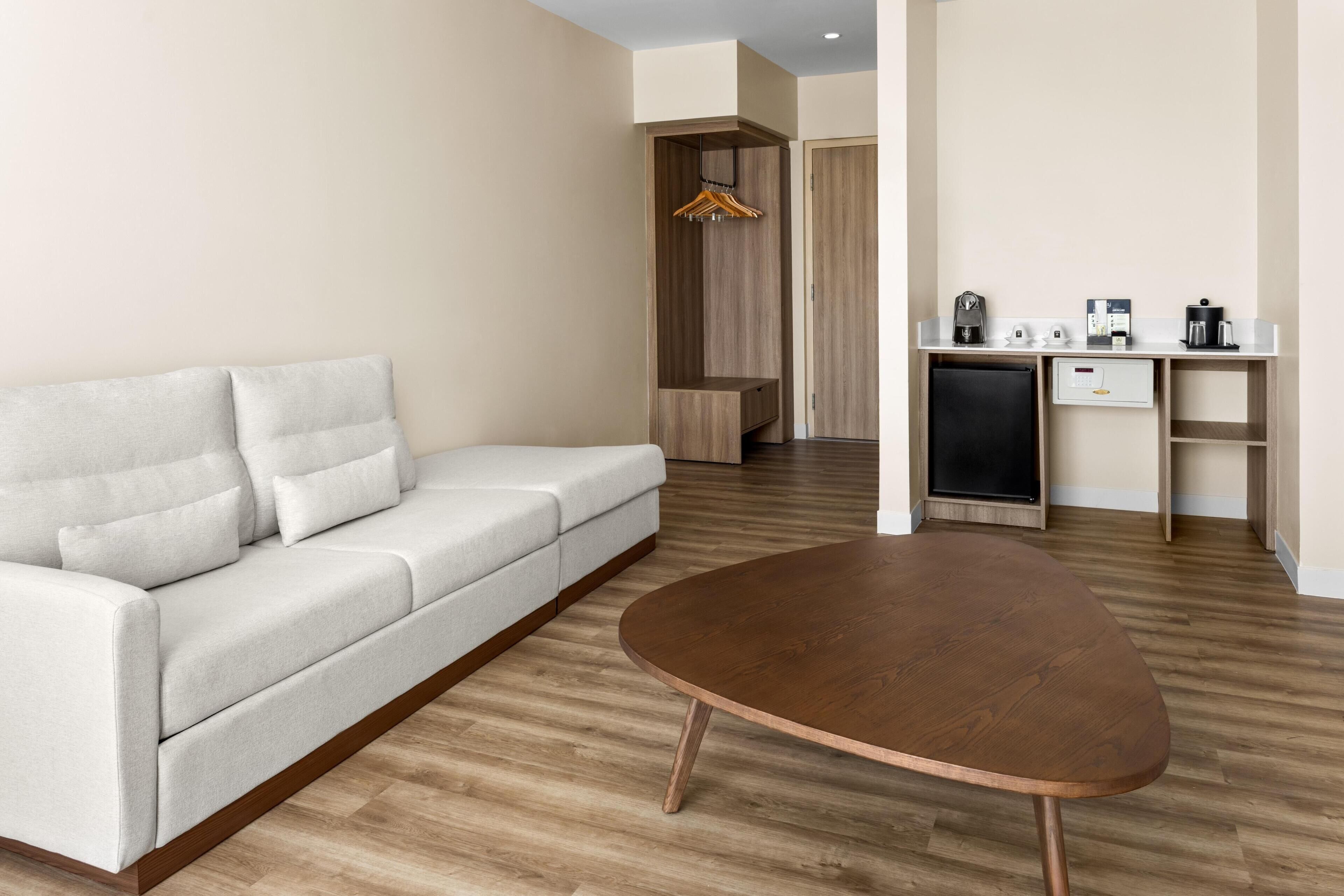 A modern living room with a sofa, a coffee table, and a kitchenette area with a refrigerator, coffee maker, and a small cabinet.