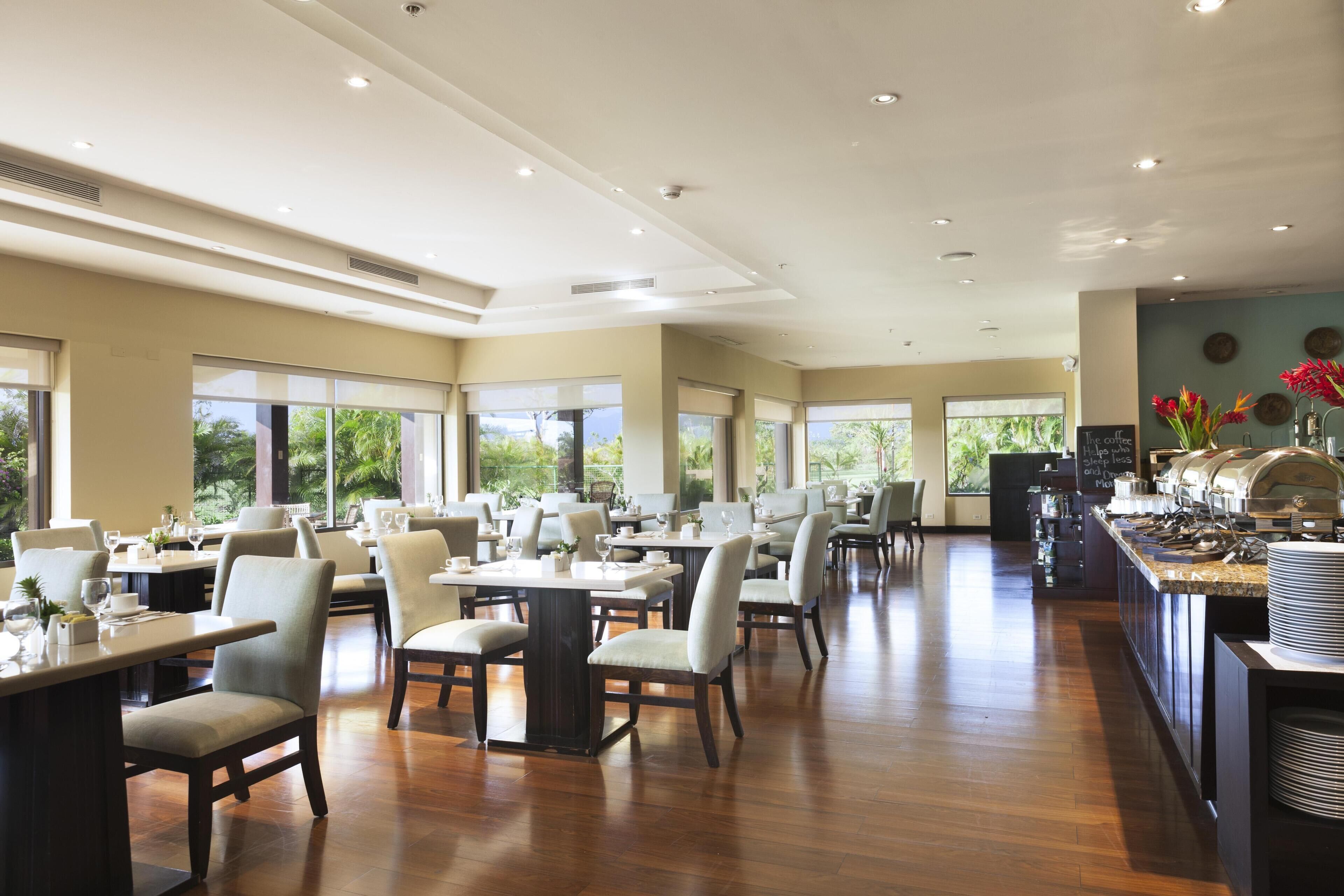 A spacious dining area with multiple tables and chairs, large windows offering views of greenery, and a buffet counter with various food items.