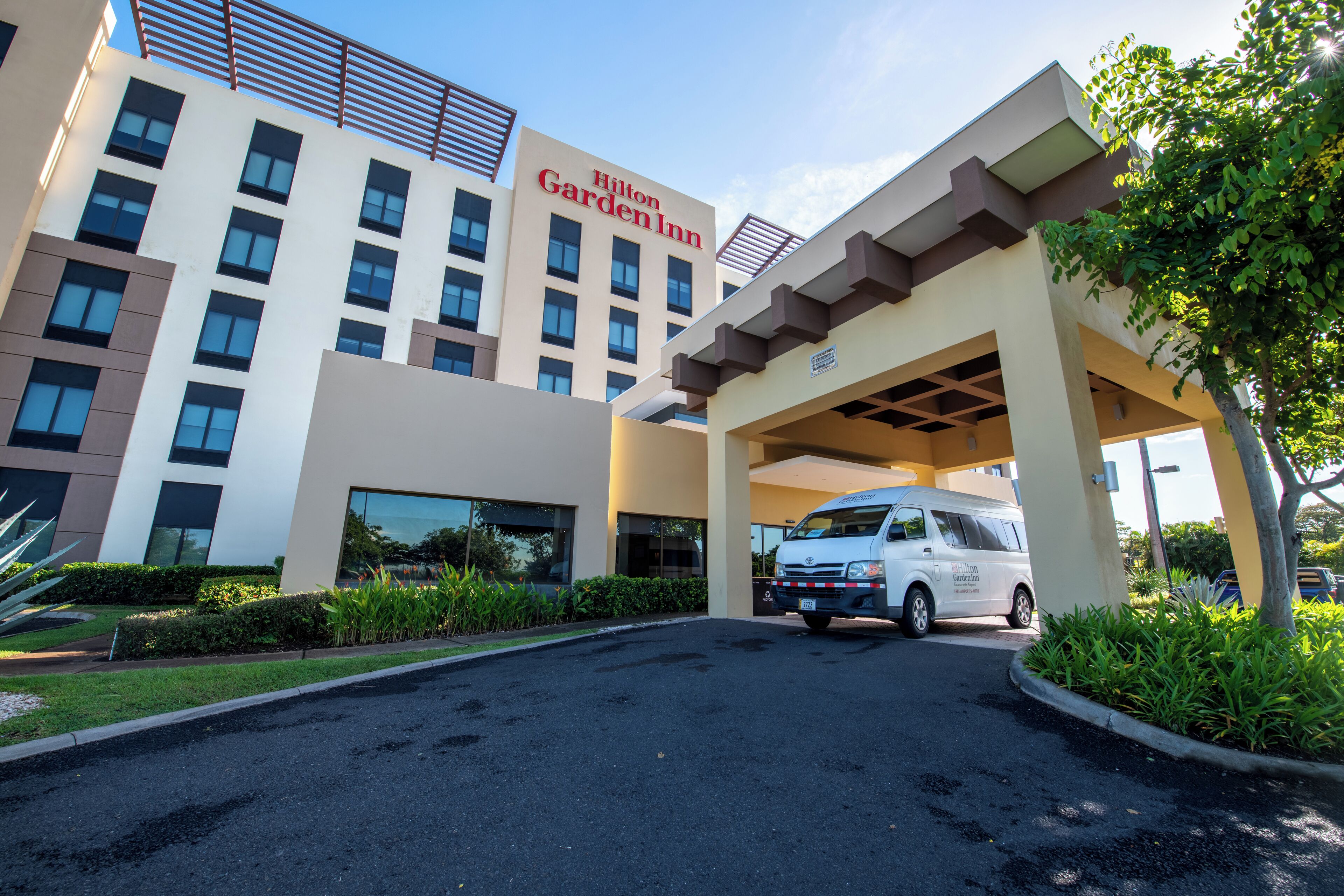 A Hilton Garden Inn with a white van parked in front, surrounded by greenery and a clear sky.