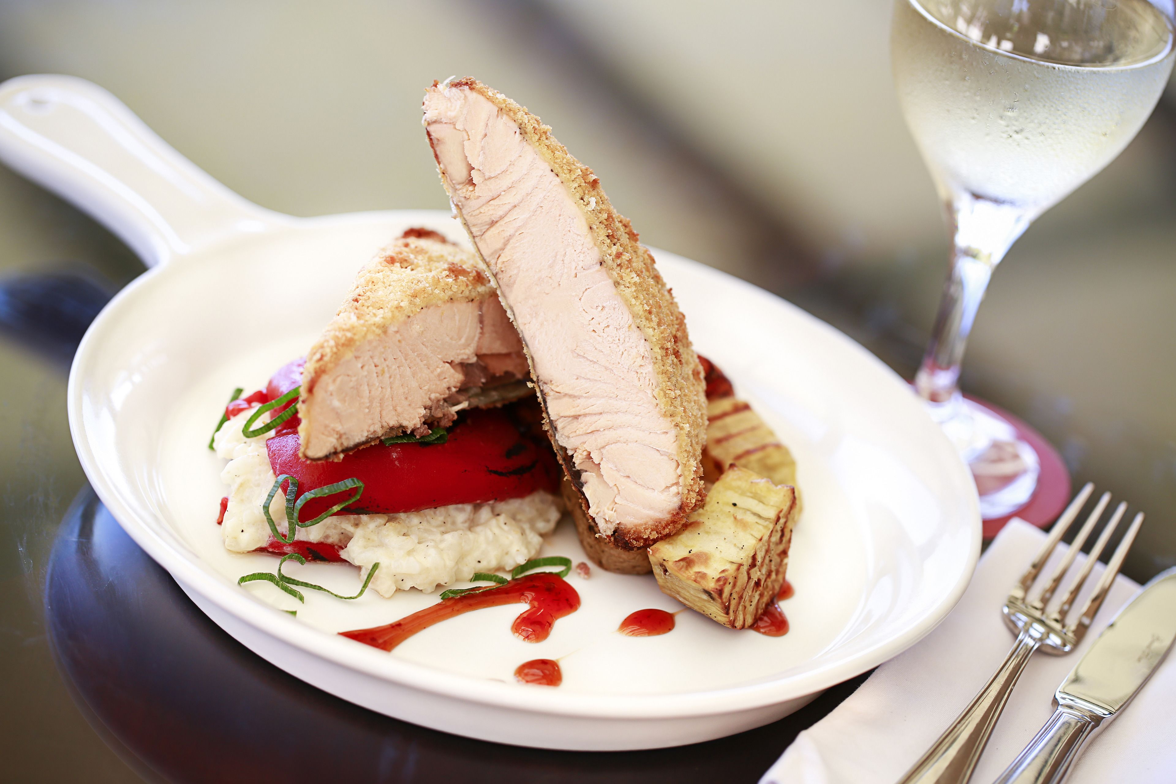 A plate with a slice of meat, vegetables, and sauce, accompanied by a glass of white wine and cutlery on a table.