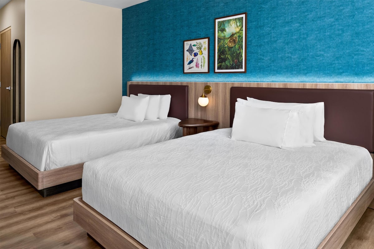 Une chambre d’hôtel avec deux lits, une tête de lit en bois, un mur d’accent bleu et des œuvres d’art encadrées.
