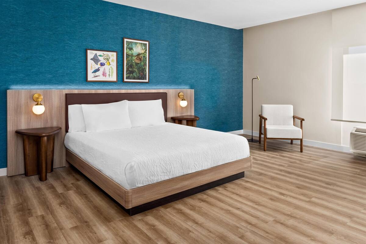 Une chambre d’hôtel moderne avec un grand lit, une tête de lit en bois, deux lampes fixées au mur, un tableau encadré, une chaise et une grande fenêtre.