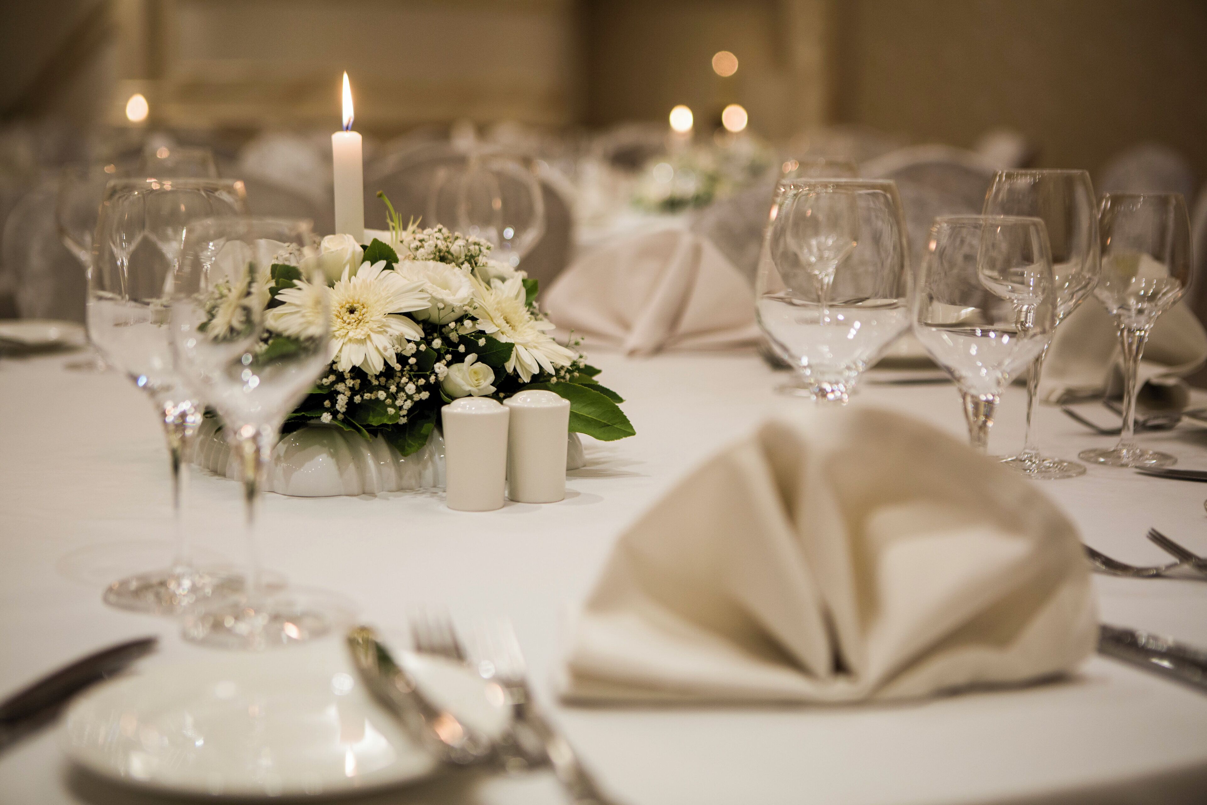 Una mesa dispuesta para un evento formal, con manteles blancos, copas de cristal transparente y un centro de mesa con flores y velas blancas.