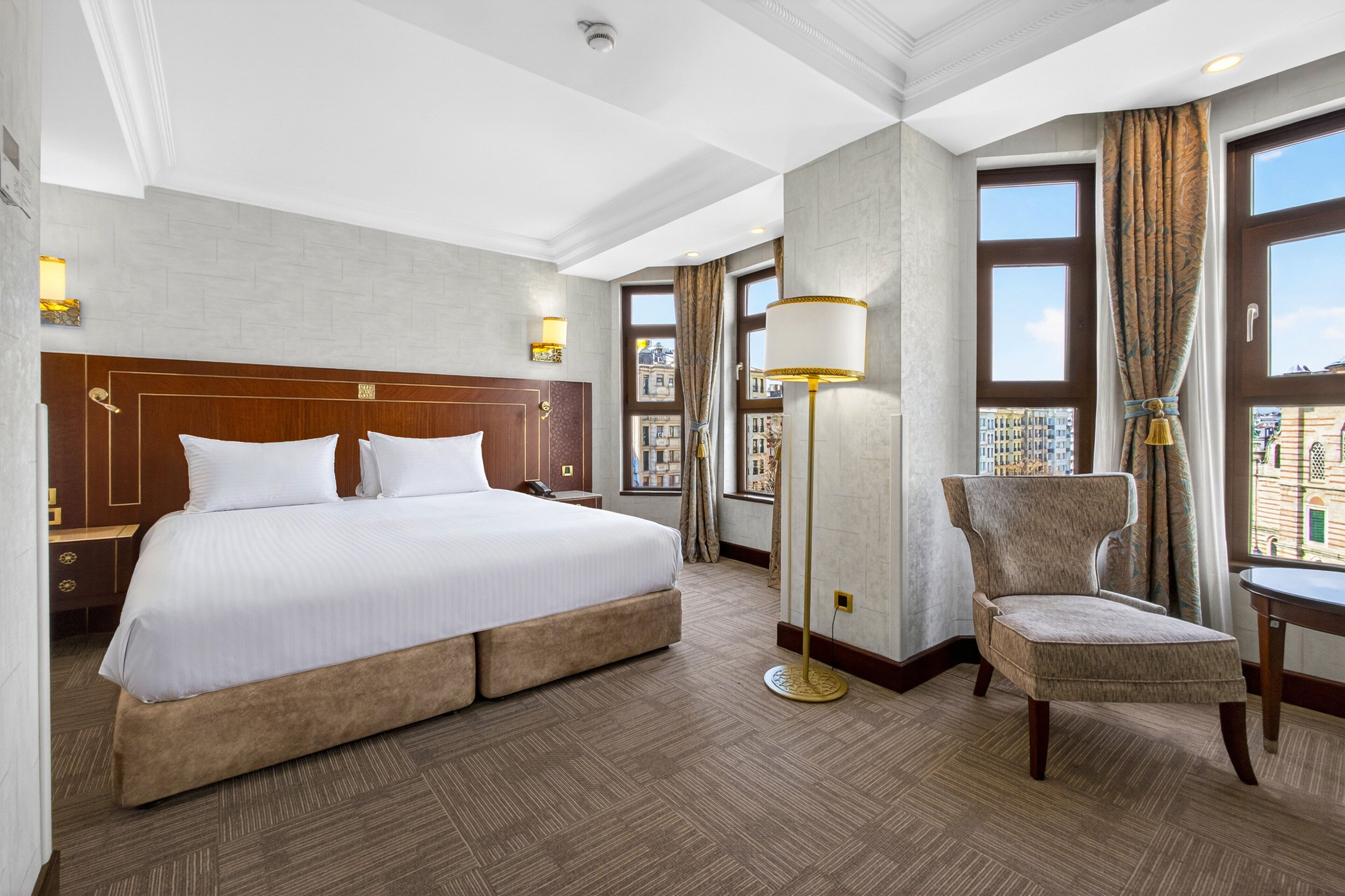 Habitación de hotel con una cama grande, una silla, una lámpara de pie y vistas a edificios a través de las ventanas.