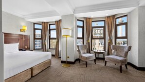 Hotelkamer met een bed, twee fauteuils, een tafeltje, een vloerlamp en grote ramen met gordijnen.