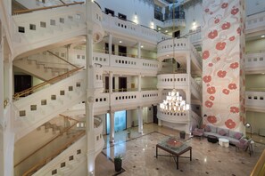 Un hall d'hôtel à plusieurs niveaux, agrémenté d'un imposant lustre, de plusieurs escaliers et d'un grand mur orné d'un motif floral.