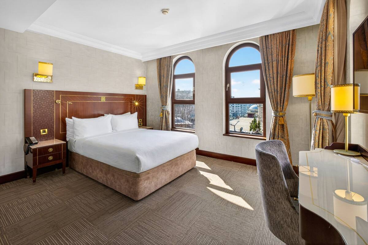 Een hotelkamer met een groot bed, een bureau, twee lampen, een nachtkastje en uitzicht op de stad.