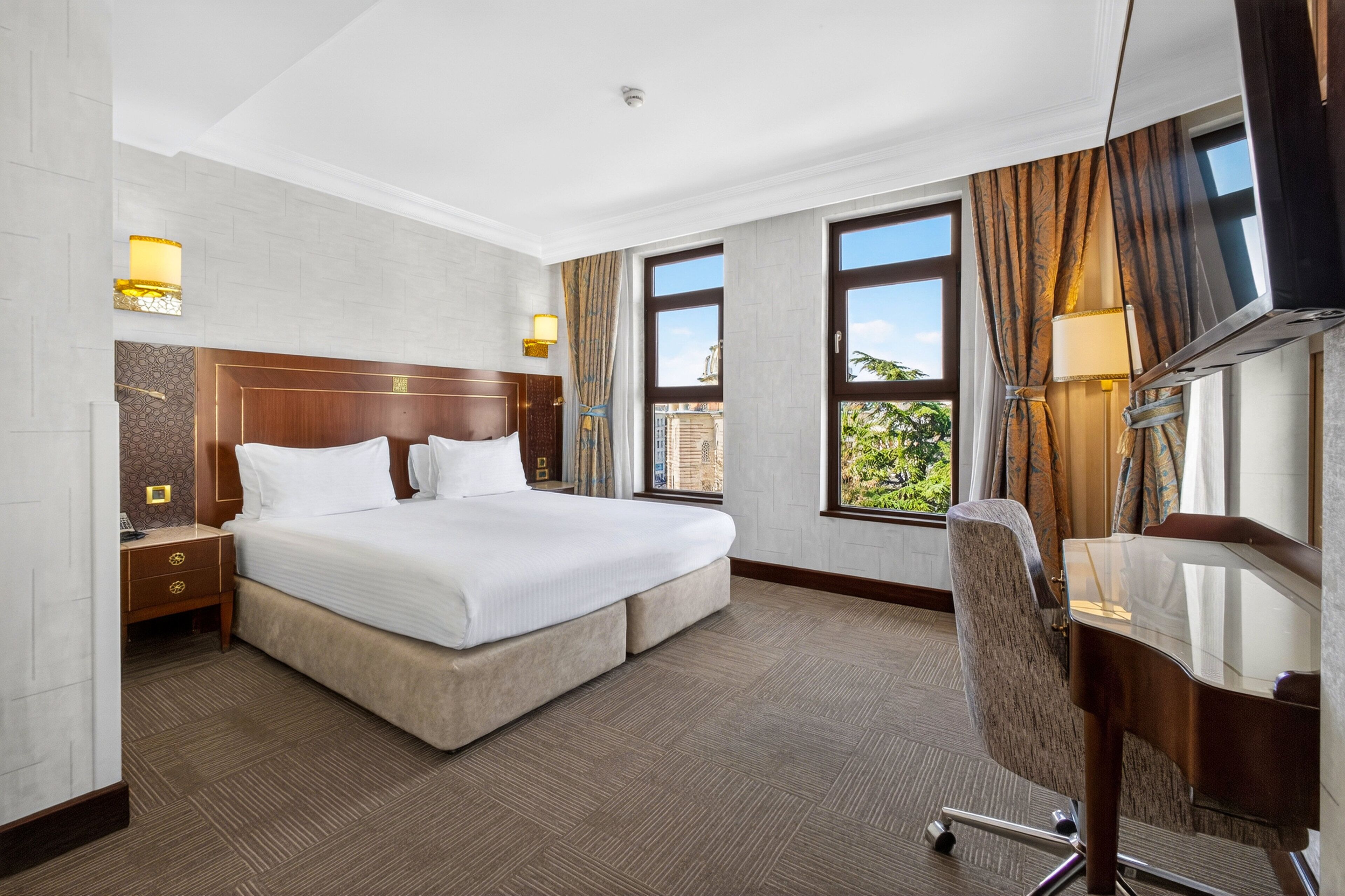 A hotel room with a large bed, a desk, a chair, and two windows with curtains.