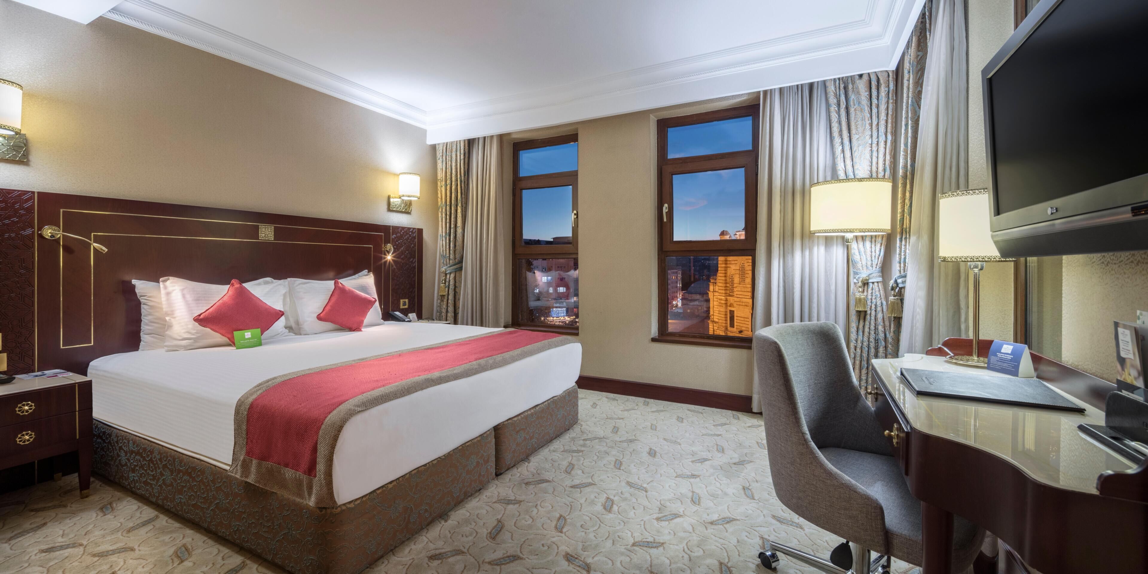 A hotel room with a large bed, a desk with a chair, a television, and a window with a city view.