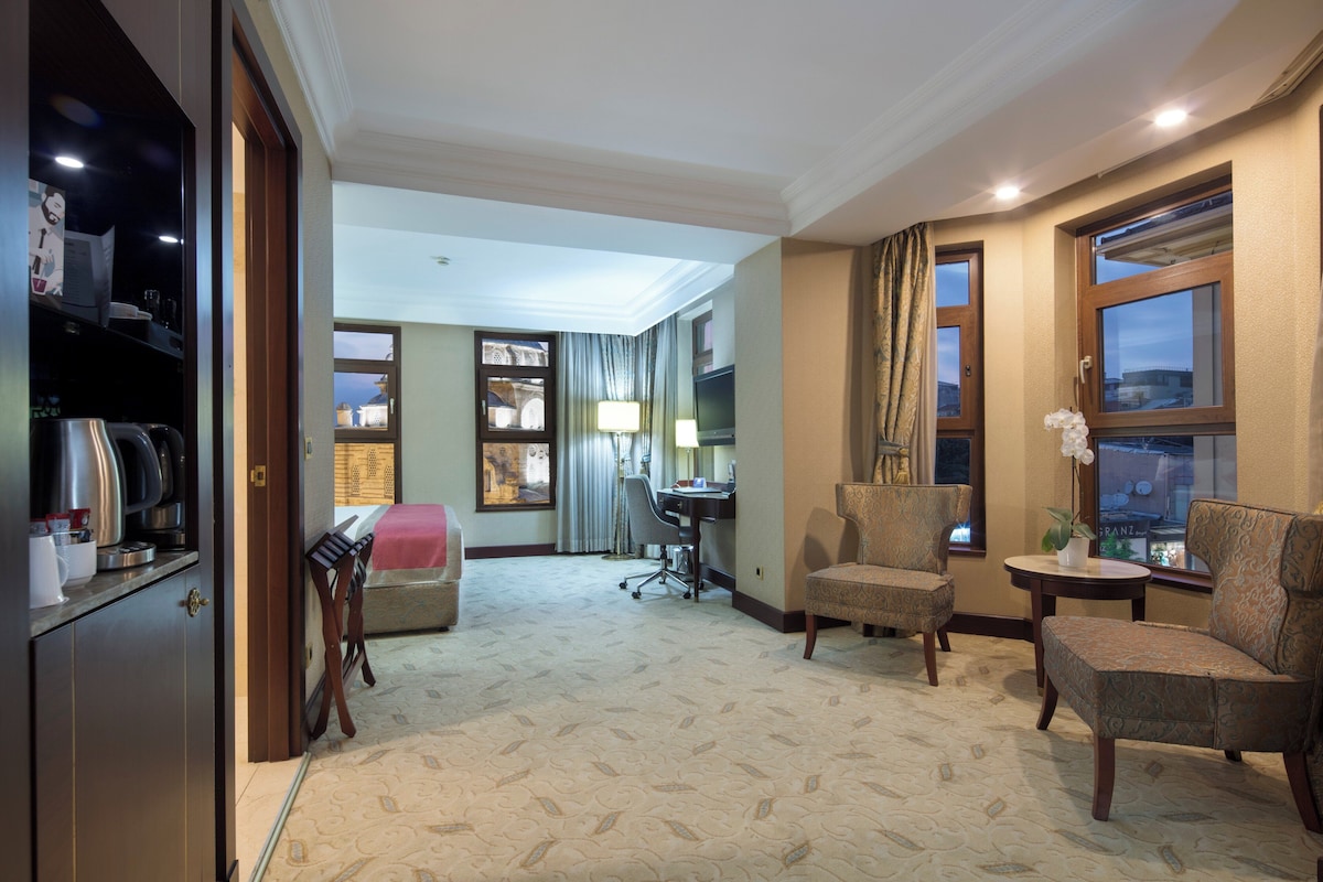 Habitación de hotel con zona de estar, escritorio, televisor y vistas a la ciudad a través de las ventanas.