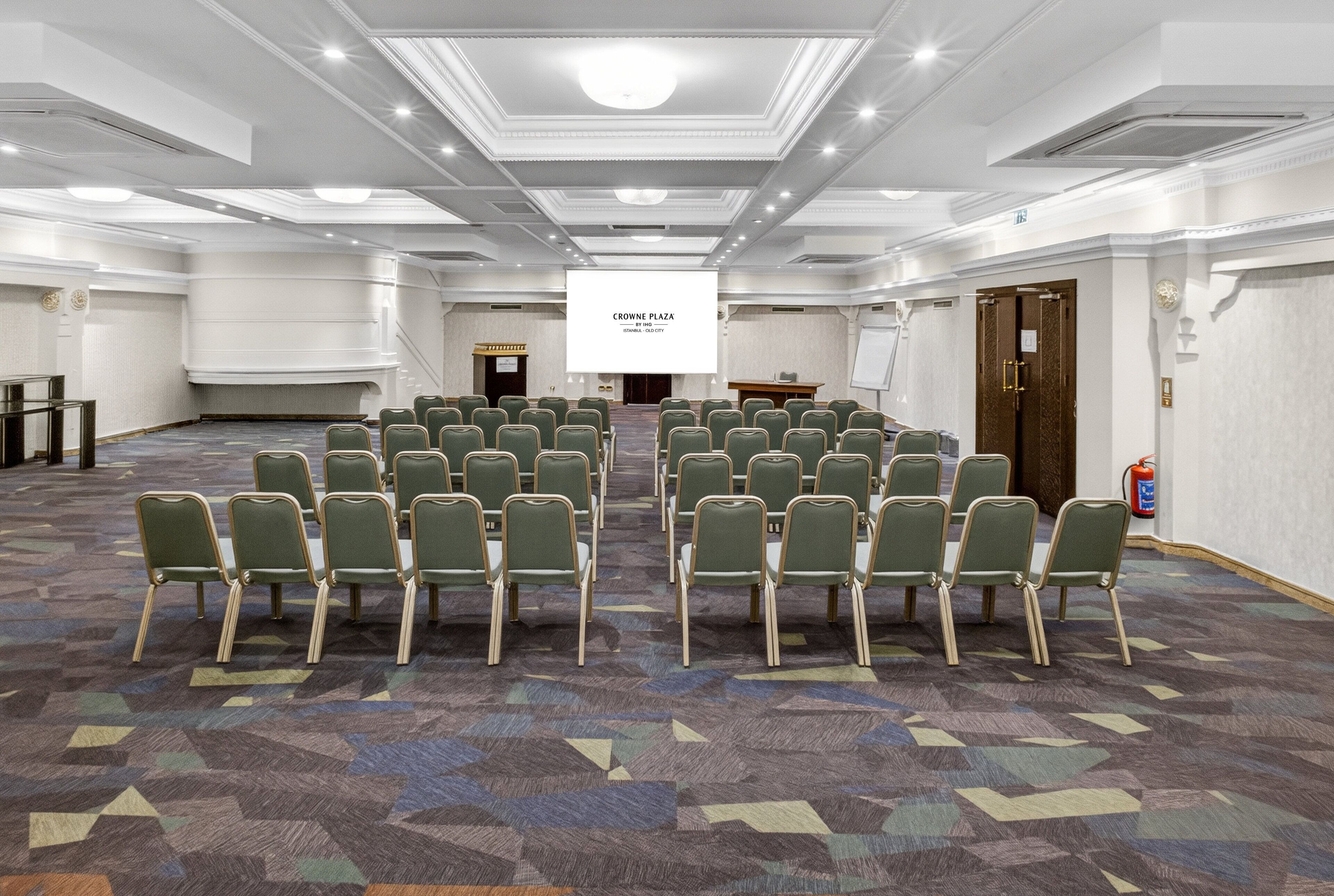 A conference room with rows of chairs, a podium, and a screen displaying 'CROWNE PLAZA.'