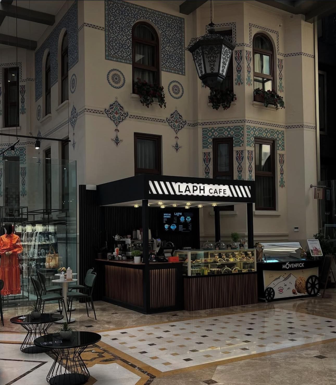 Ein Café namens LAPH CAFE mit modernem Design, das eine Theke mit Speisekarte, einen Sitzbereich mit Tischen und Stühlen sowie dekorative Wandfliesen bietet.