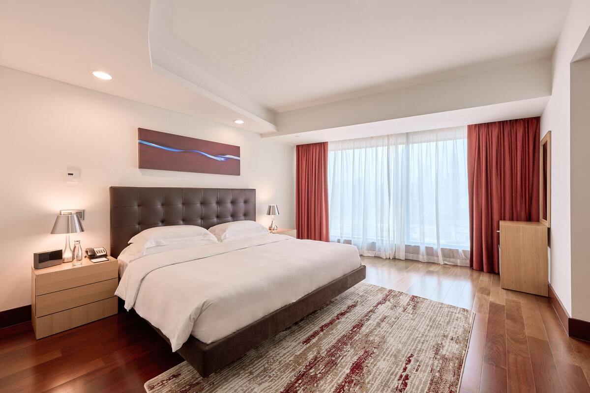 A modern bedroom with a large bed, wooden nightstands, a bedside table with a lamp, a large window with curtains, and a wall-mounted artwork.