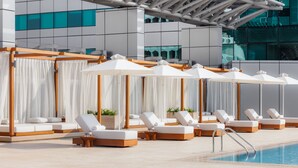 Zona de piscina con sillones de descanso y sombrillas, una piscina y un edificio moderno al fondo.
