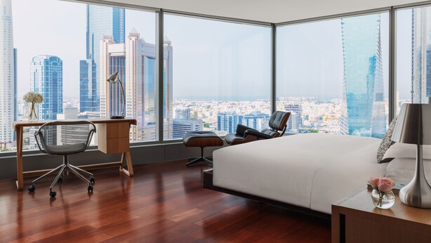 Una habitación de hotel moderna con una cama grande, un escritorio, una silla y vistas a un paisaje urbano a través de grandes ventanales.