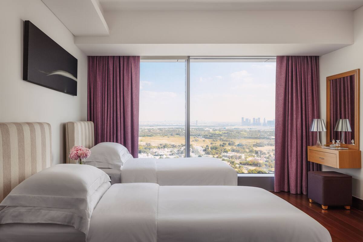 A hotel room with two beds, a large window with curtains, a city view, a wooden dresser, and a painting on the wall.
