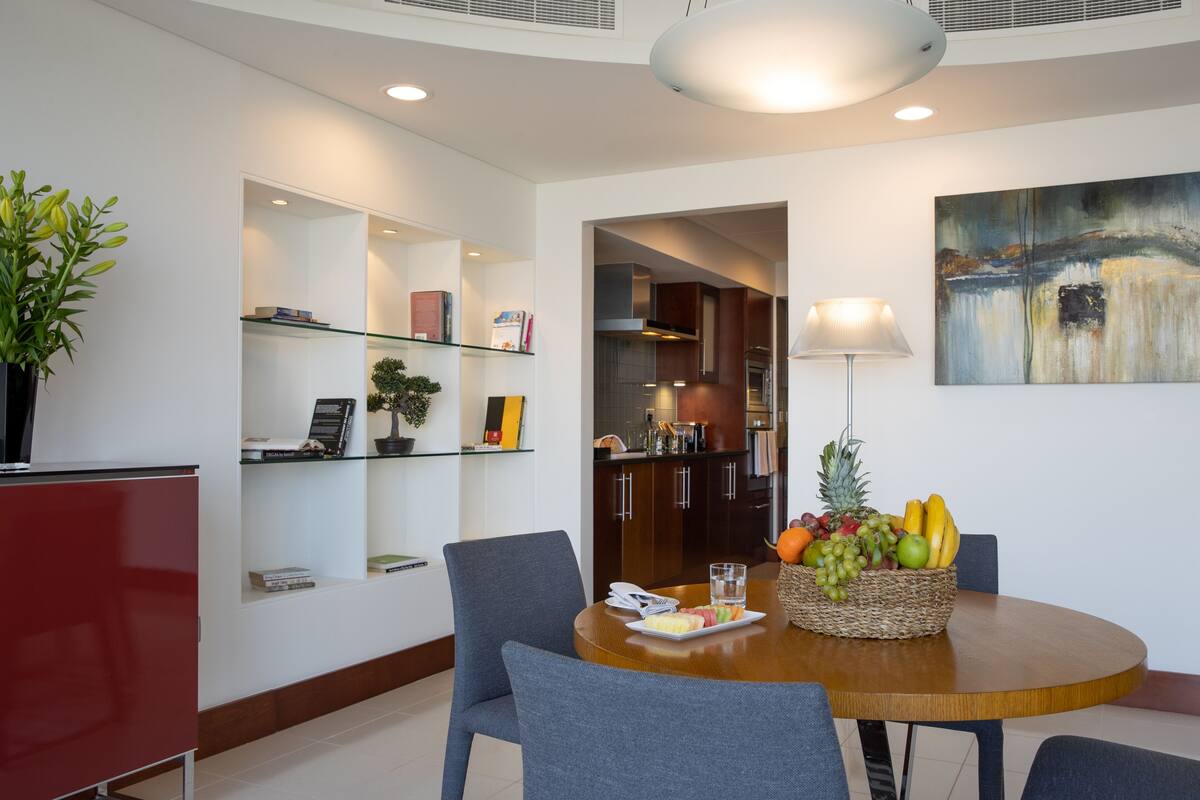 A modern dining area with a round wooden table, grey chairs, a fruit basket, and a glass shelf with books and decorative items.