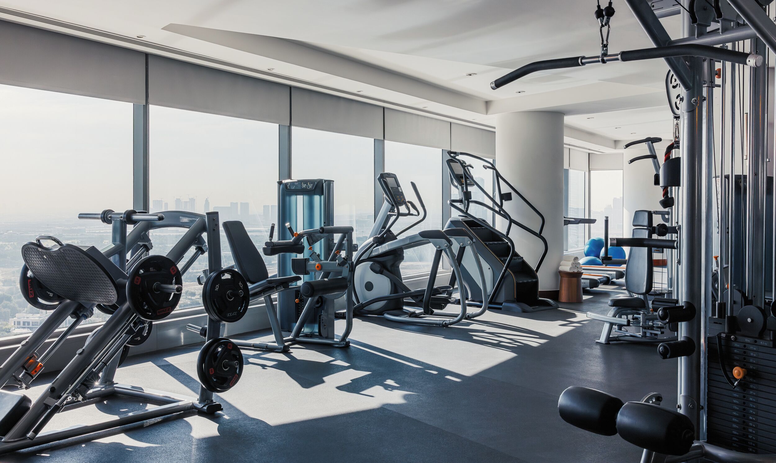 A modern gym with various exercise equipment, large windows, and a city view.