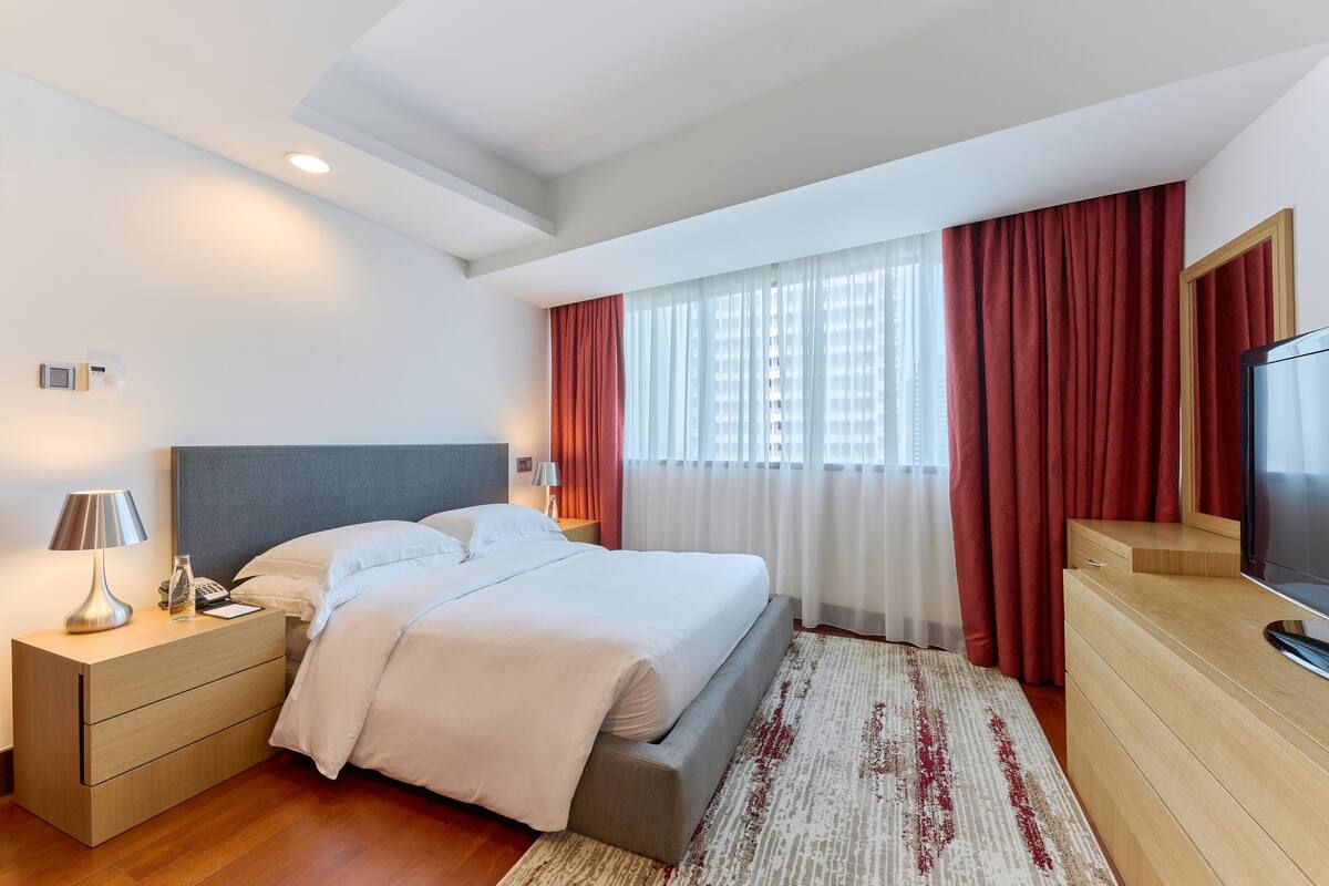 A modern hotel room with a bed, bedside tables, a TV, and a large window with curtains.