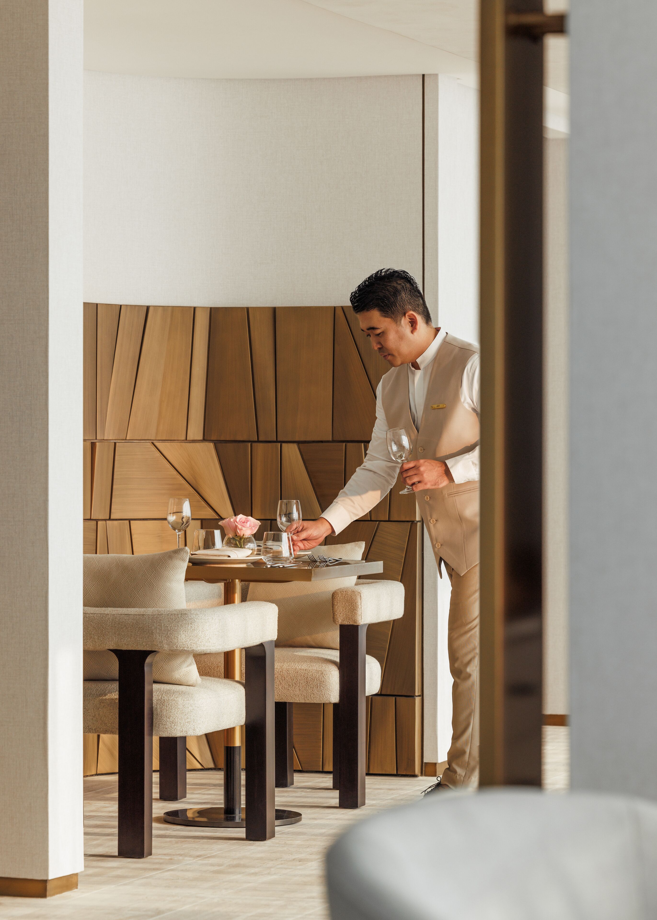 A modern dining area with a wooden wall panel, a table set with a rose, and a person in a beige suit.