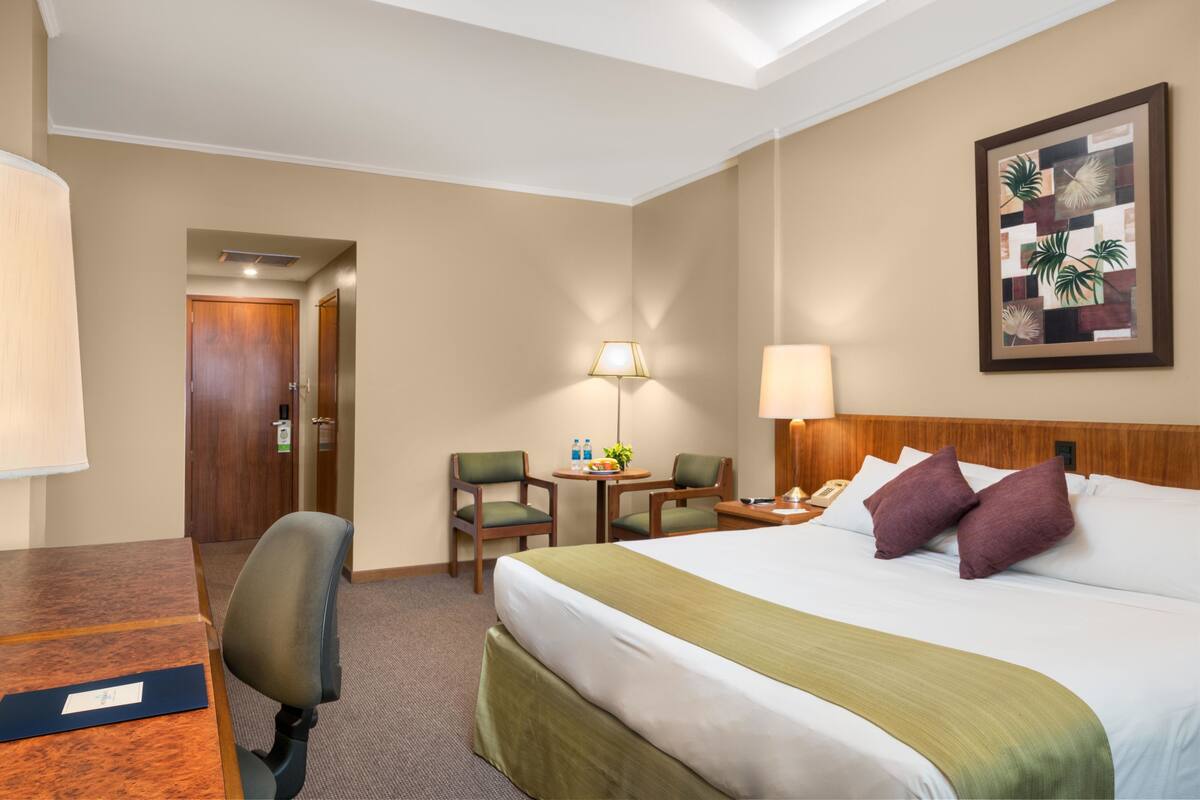 Une chambre d’hôtel avec un lit, deux chaises, un bureau, une lampe et un tableau encadré au mur.
