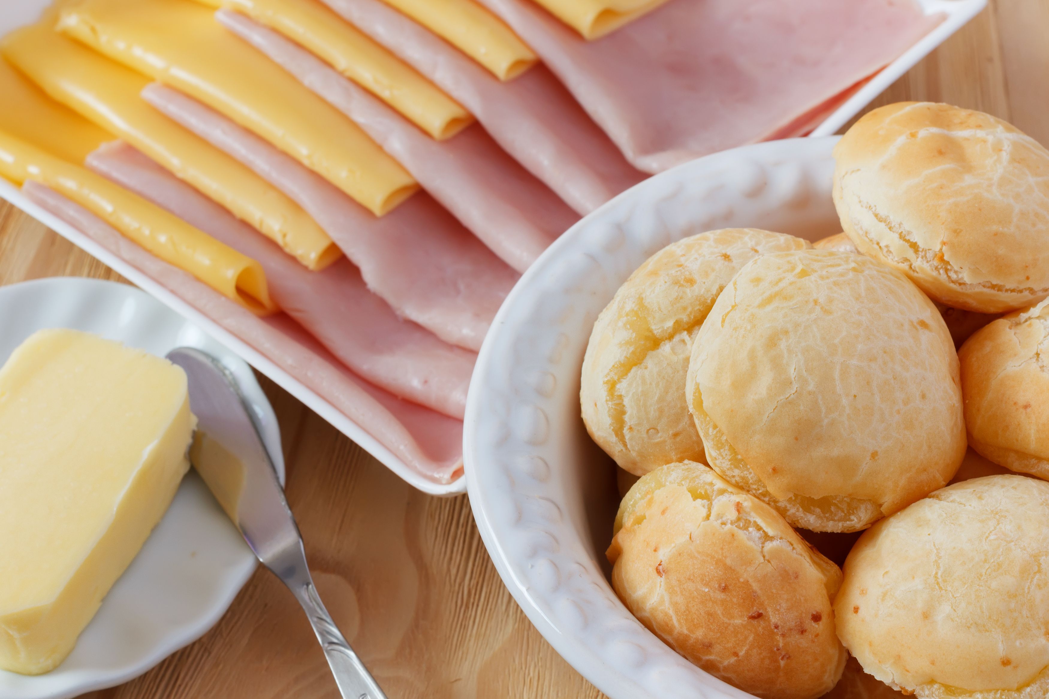 A plate with sliced cheese and ham, a bowl of bread rolls, and a block of butter on a wooden table.