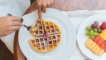 A plate of waffles with syrup and powdered sugar, a plate of fresh fruit, and a cup of coffee on a wooden table.
