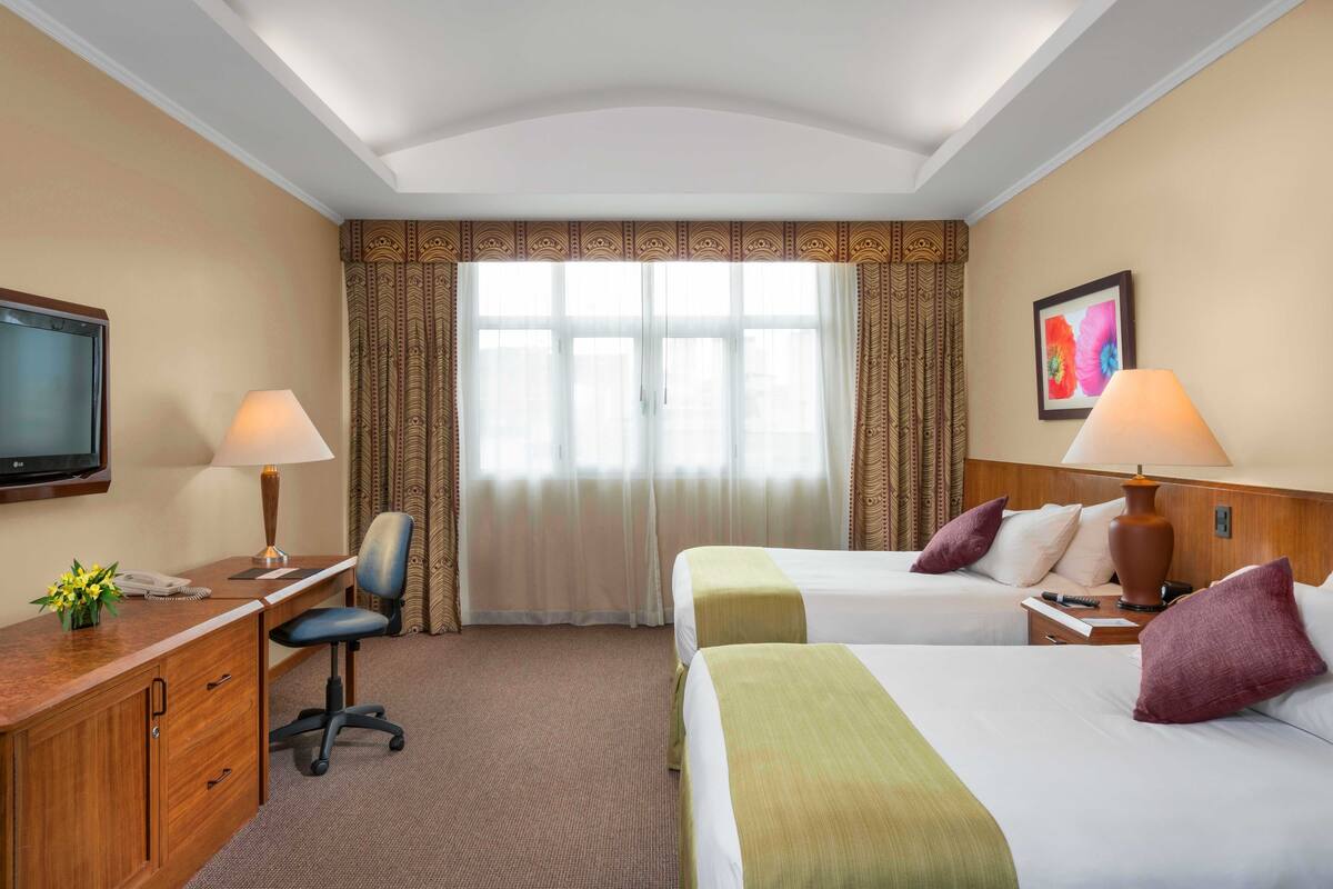 Une chambre d’hôtel avec deux lits, un bureau avec une chaise, une télévision et une fenêtre avec des rideaux.