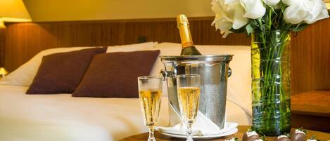 A hotel room with a bed, a bedside table with champagne, glasses, and chocolates.