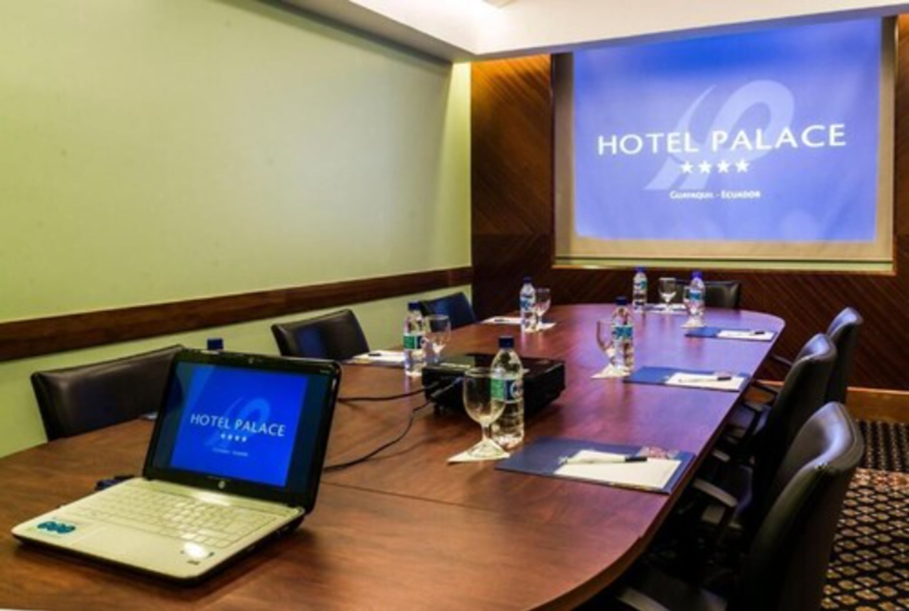 Ein Konferenzraum mit einem Laptop, Wasserflaschen und einer Projektionsleinwand, die "HOTEL PALACE" anzeigt.