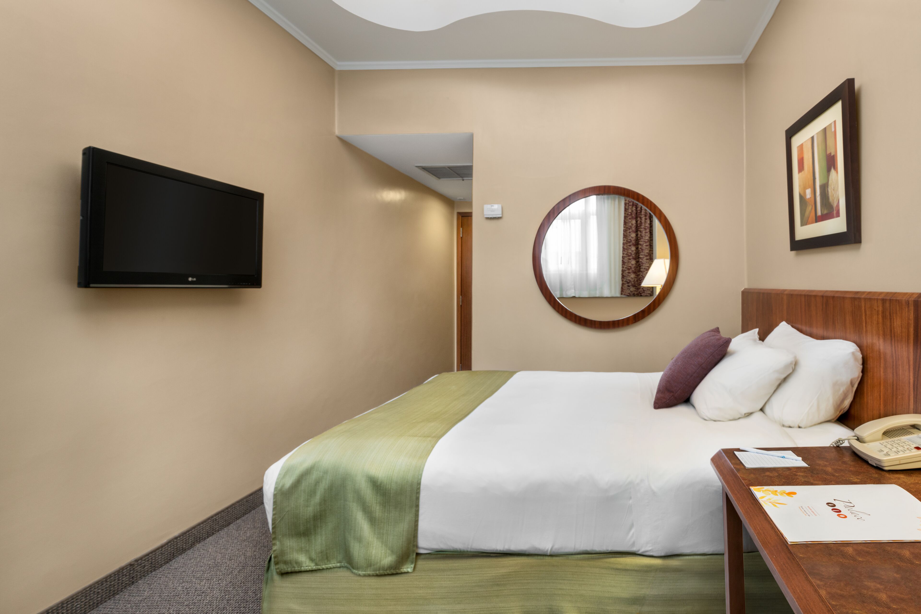 Habitación de hotel con cama, mesita de noche con teléfono, televisor de pantalla plana en la pared y espejo.