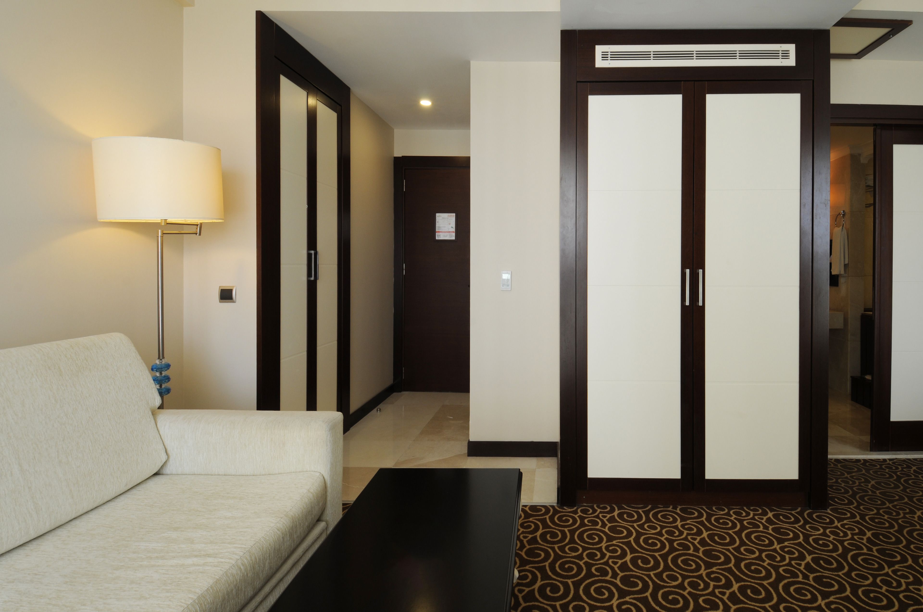 A hotel room with a sofa, a lamp, a patterned carpet, and a wardrobe with a mirror.