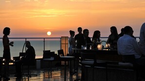 Een zonsondergang uitzicht vanaf een rooftop bar, met mensen zittend en staand, met uitzicht op de oceaan.