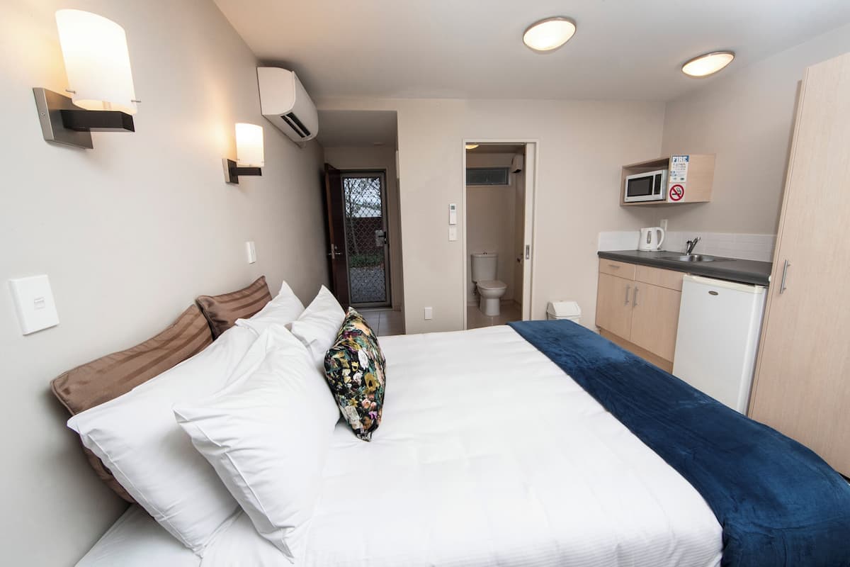 A bedroom with a bed, pillows, a bedside table, a microwave, and a small kitchenette.