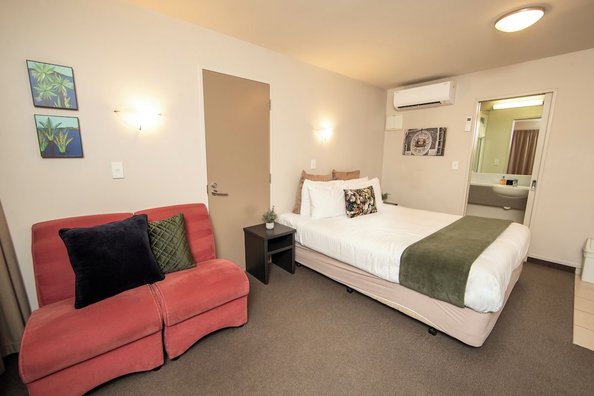 A hotel room with a bed, a red sofa, a nightstand, and wall-mounted artwork.