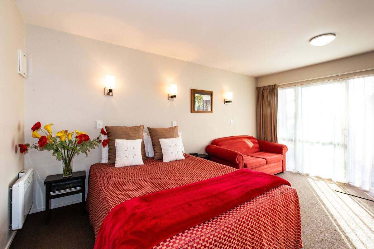 A hotel room with a large bed, a red armchair, a vase of flowers, and a window with curtains.