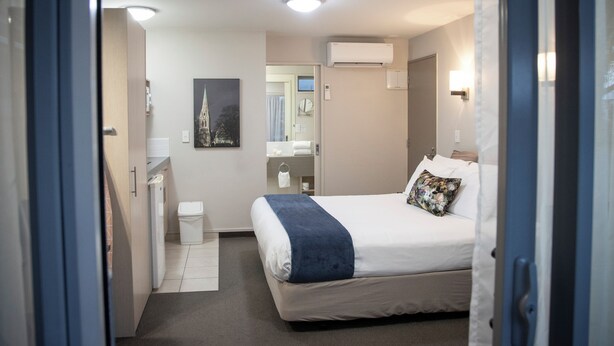 Ett hotellrum med en säng, ett litet sängbord, en spegel, en luftkonditioneringsanläggning och en dörr som leder till ett annat rum.