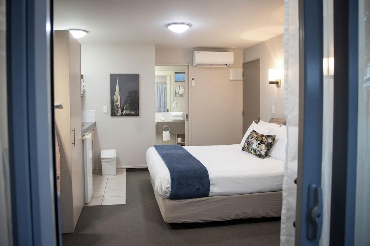 A hotel room with a bed, a small bedside table, a mirror, an air conditioning unit, and a door leading to another room.