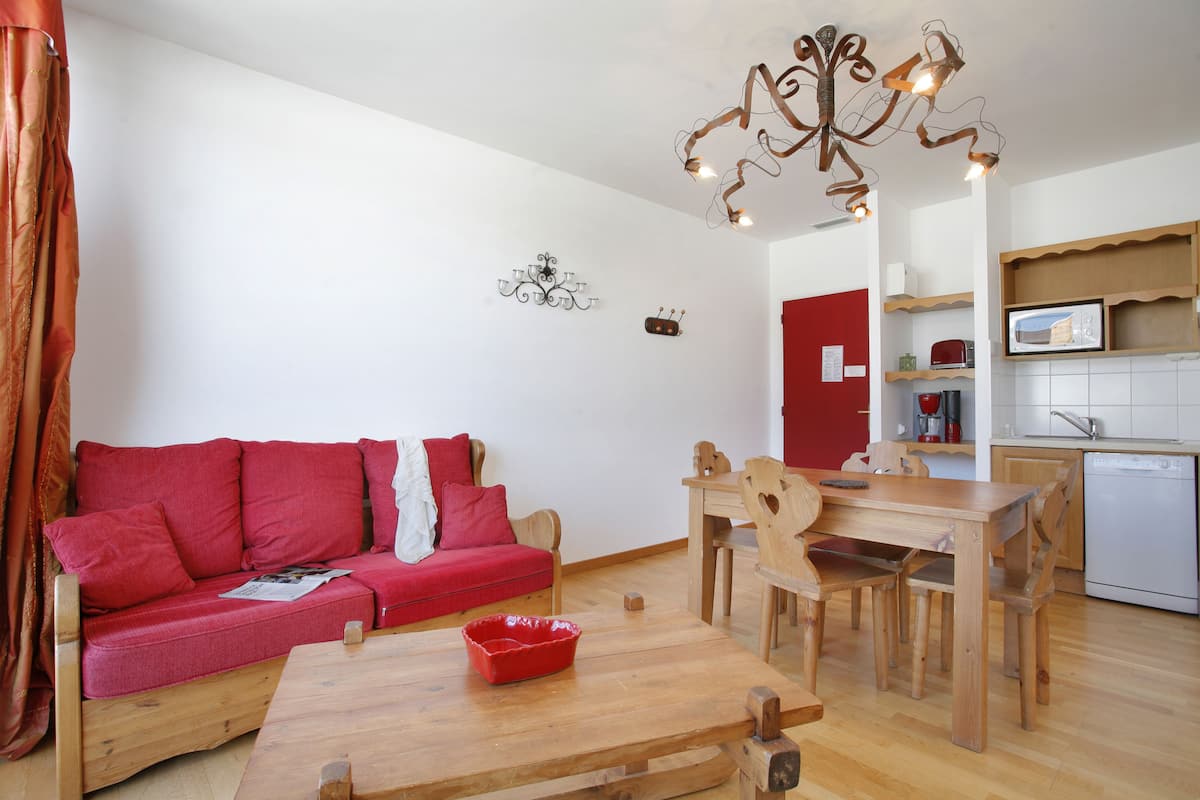 Un espacio habitable compacto con sofá rojo, mesa de comedor de madera y una zona de cocina con microondas.