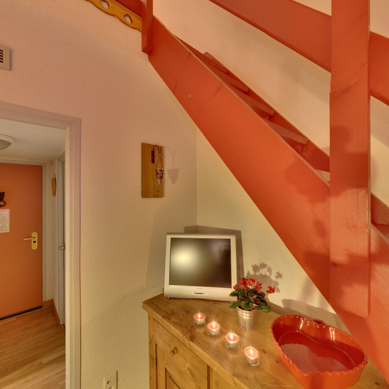 Una escalera con barandilla de madera y una pared blanca. Un televisor está montado en la pared, y hay velas sobre una mesa de madera.