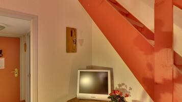 Una escalera con barandilla de madera y una pared blanca. Un televisor está montado en la pared, y hay velas sobre una mesa de madera.