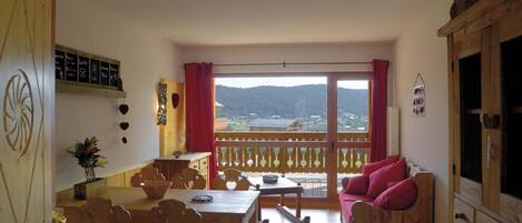 A cozy room with a wooden dining table, chairs, a sofa with red cushions, and a balcony with a view of the countryside.