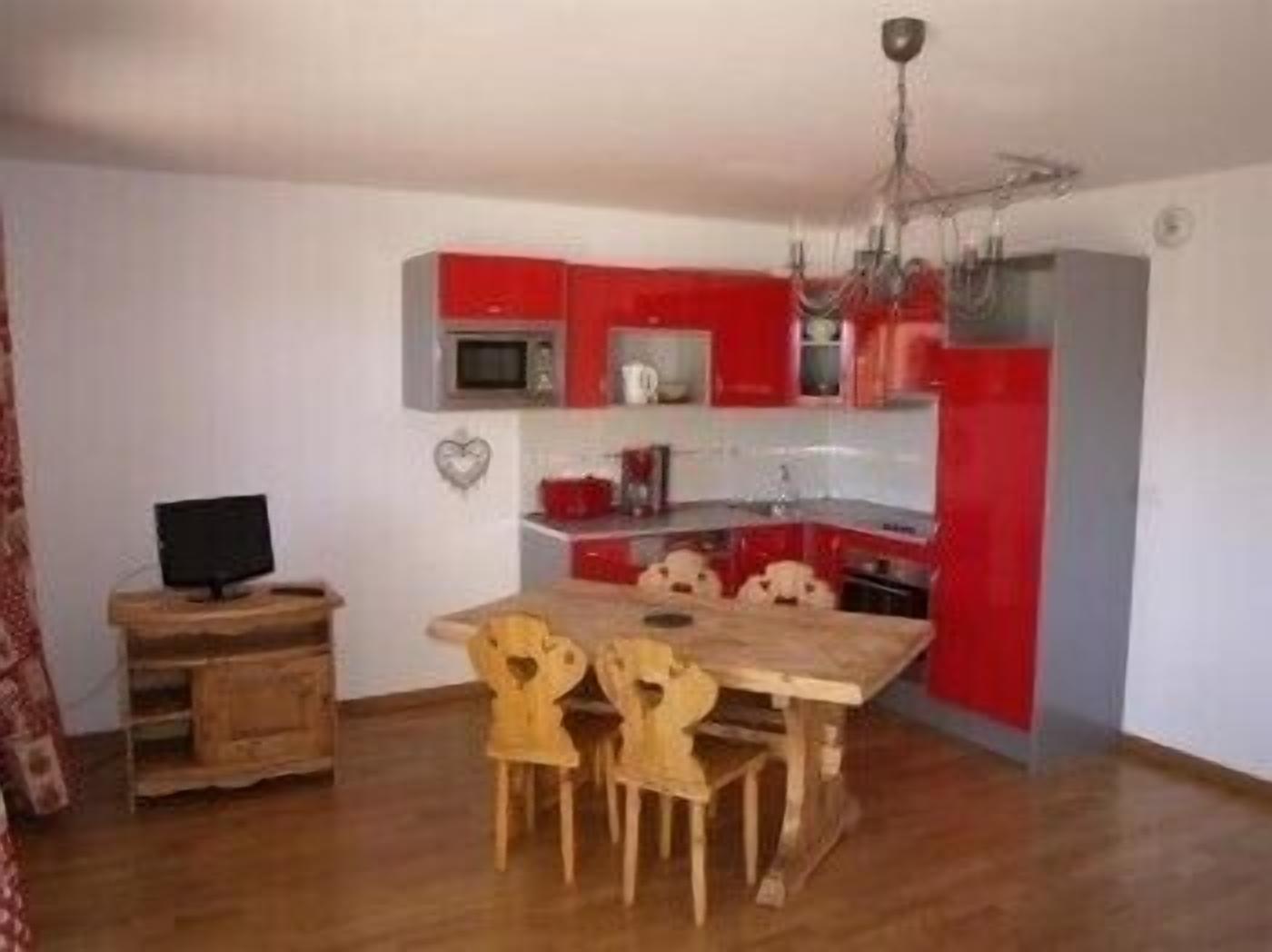 Una cocina compacta con armarios rojos, una mesa de comedor de madera y un televisor sobre una mesita auxiliar.