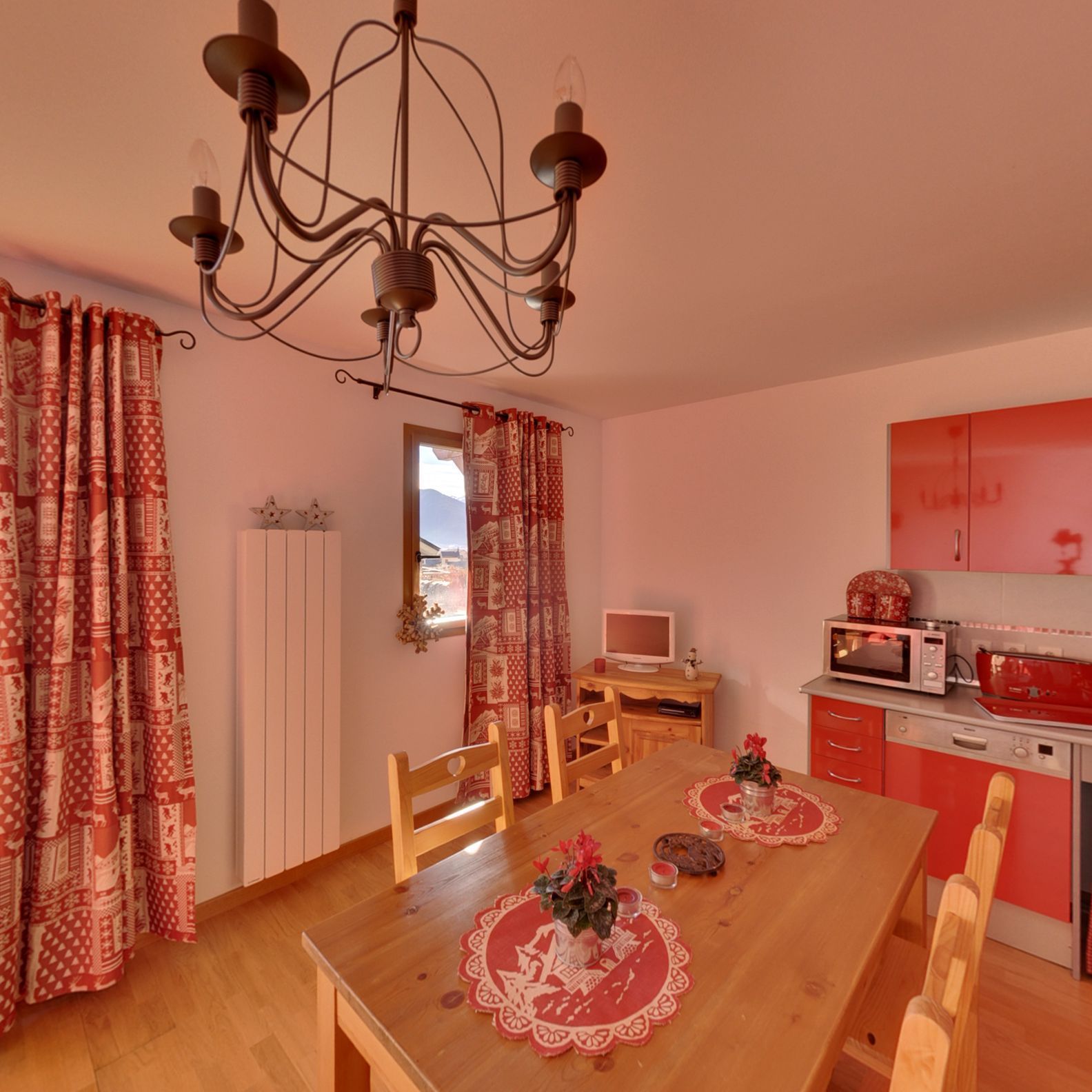 A cozy dining area with a wooden table, chairs, and a chandelier. A kitchen with red cabinets and appliances is visible in the background.