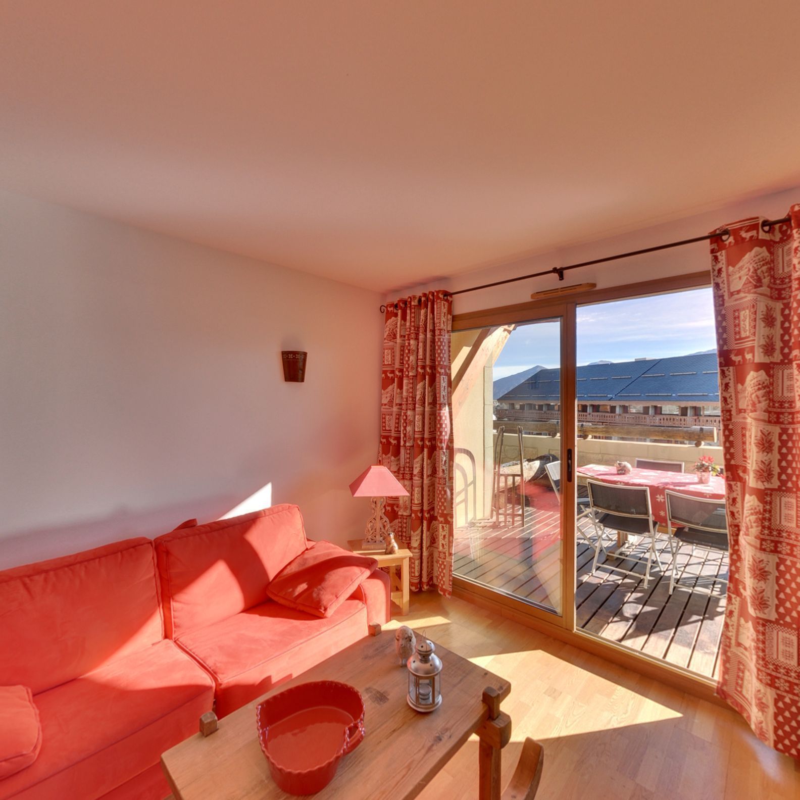 Una habitación con sofá rojo, una mesa de madera con un cubo rojo, una lámpara y vistas a un balcón con mobiliario exterior.