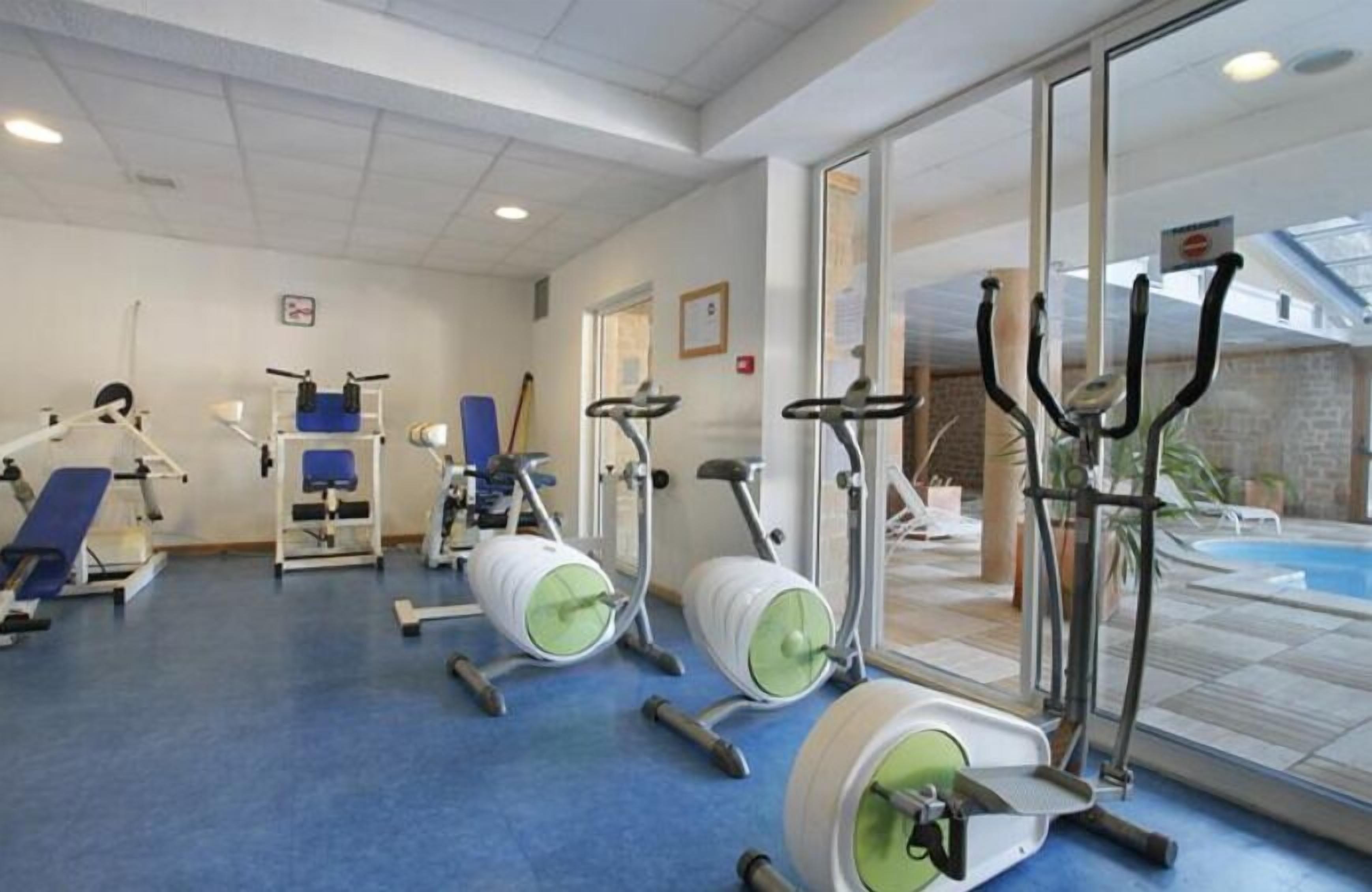 A gym with various exercise equipment, a blue floor, and a pool visible through the glass doors.