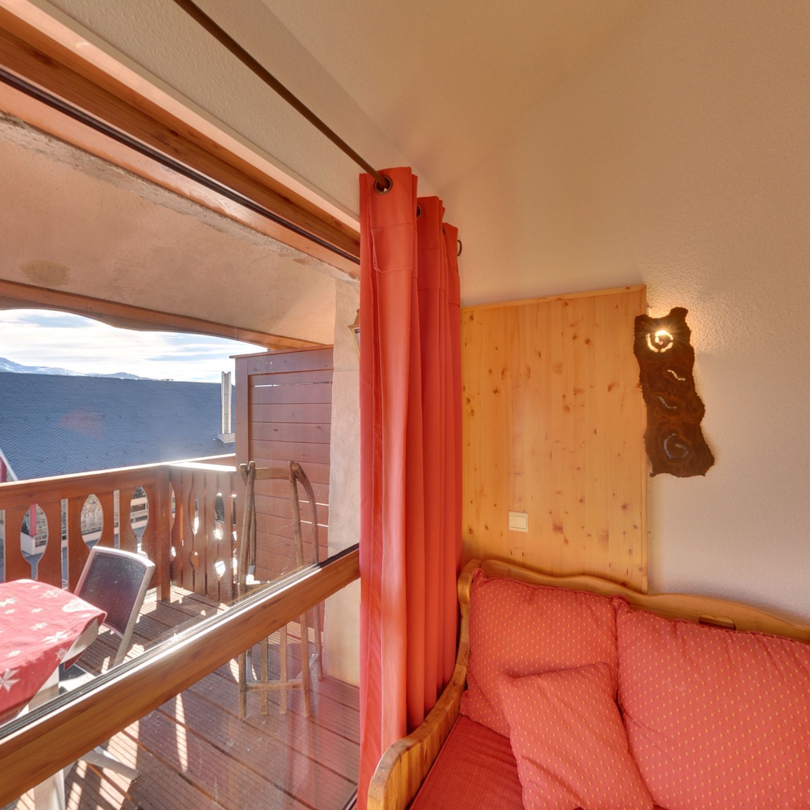 A room with a wooden headboard, a bed with a red polka dot cover, a wooden deck with a table and chairs, and a view of the mountains.